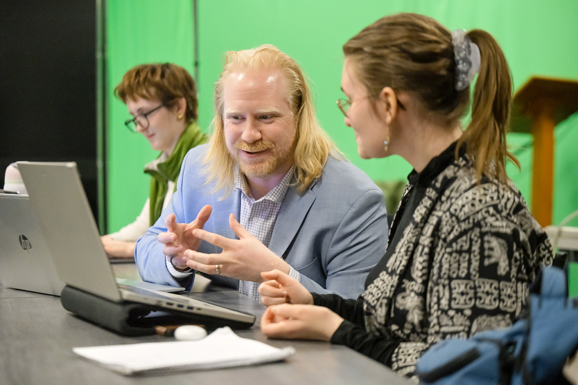 Smiling individual helps student with another student visible in background.